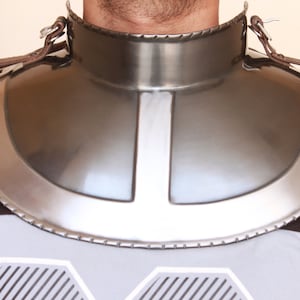 Könnte beinhalten: Ein silberner Nackenschutz aus Metall mit einem braunen Lederriemen. Der Nackenschutz ist gebogen und hat ein dekoratives Design.