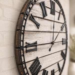 K&ouml;nnte beinhalten: Eine gro&szlig;e, runde Wanduhr mit rustikalem Design. Die Uhr hat ein wei&szlig;es Holzgesicht mit schwarzen r&ouml;mischen Ziffern und Zeigern. Der &auml;u&szlig;ere Rahmen ist dunkelbraun, und die Uhr ist an einer wei&szlig;en Backsteinmauer befestigt.