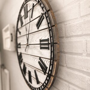 K&ouml;nnte beinhalten: Gro&szlig;e, runde Wanduhr mit einem rustikalen, antiken Design. Das Zifferblatt ist wei&szlig; mit schwarzen r&ouml;mischen Ziffern und Zeigern. Der Rahmen besteht aus Holz mit einer verwitterten, braunen Oberfl&auml;che. Die Uhr ist an einer wei&szlig;en Backsteinmauer befestigt.