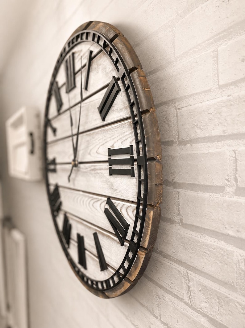 K&ouml;nnte beinhalten: Gro&szlig;e, runde Wanduhr mit einem rustikalen, antiken Design. Das Zifferblatt ist wei&szlig; mit schwarzen r&ouml;mischen Ziffern und Zeigern. Der Rahmen besteht aus Holz mit einer verwitterten, braunen Oberfl&auml;che. Die Uhr ist an einer wei&szlig;en Backsteinmauer befestigt.