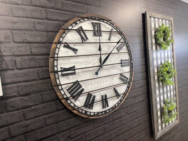 K&ouml;nnte beinhalten: Gro&szlig;e, runde Wanduhr mit einem wei&szlig;en Holzgesicht im Used-Look und schwarzen r&ouml;mischen Ziffern. Die Uhr hat einen dunkelbraunen Holzrahmen und schwarze Zeiger. Die Uhr ist an einer dunkelgrauen Backsteinmauer befestigt.
