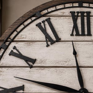 K&ouml;nnte beinhalten: Nahaufnahme einer gro&szlig;en, dekorativen Wanduhr. Das Zifferblatt hat eine wei&szlig;e Oberfl&auml;che mit schwarzen r&ouml;mischen Ziffern und Zeigern. Der Rahmen ist aus dunkelbraunem Holz mit schwarzem Metallbesatz. Die Zeiger zeigen fast auf 12 Uhr.