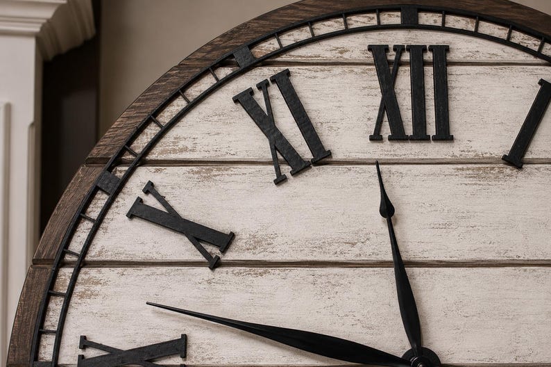 K&ouml;nnte beinhalten: Nahaufnahme einer gro&szlig;en, dekorativen Wanduhr. Das Zifferblatt hat eine wei&szlig;e Oberfl&auml;che mit schwarzen r&ouml;mischen Ziffern und Zeigern. Der Rahmen ist aus dunkelbraunem Holz mit schwarzem Metallbesatz. Die Zeiger zeigen fast auf 12 Uhr.
