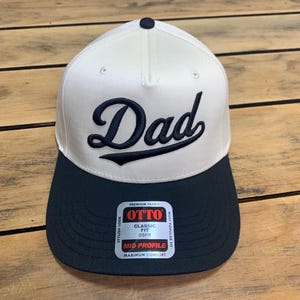Könnte beinhalten: Eine zweifarbige Baseballkappe mit weißer Krone und schwarzem Schirm. Das Wort Dad ist in schwarzer Schreibschrift auf die Vorderseite gestickt. Ein Aufkleber auf dem Schirm zeigt OTTO PREMIUM FABRIC CLASSIC FIT OSFM MID PROFILE STYLISH LOOK MOST POPULAR FIT.