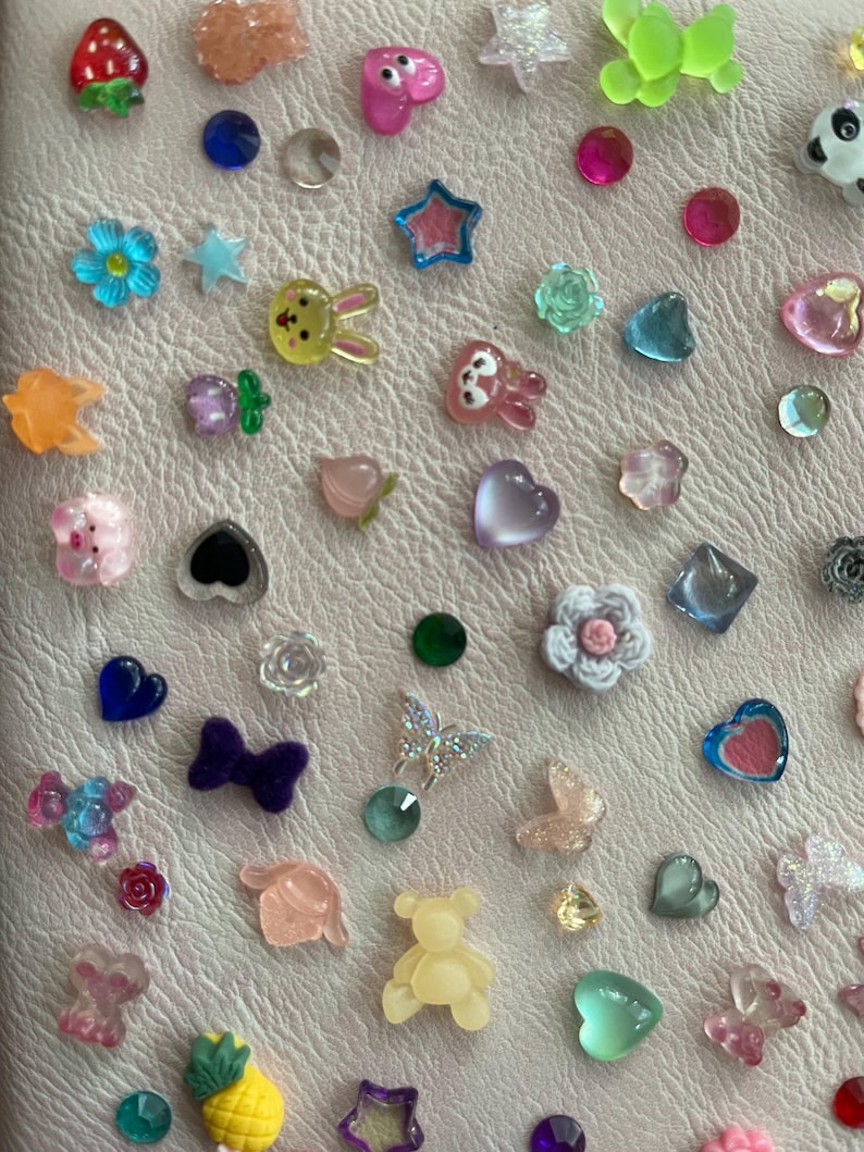 May include: A collection of colorful, small, decorative embellishments. The assortment includes various shapes like hearts, stars, flowers, butterflies, and animals, in shades of pink, blue, green, yellow, and clear. These are likely for crafts or nail art.