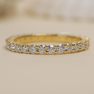 May include: A gold band ring with a continuous row of round, clear gemstones. The ring is set against a soft, neutral background, highlighting the sparkle of the stones. The ring is a classic design.