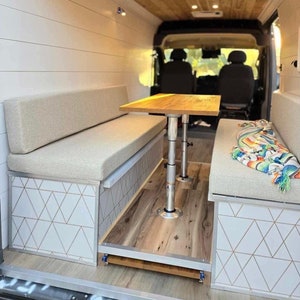 May include: Interior view of a van conversion featuring a wooden table and benches with light-colored cushions. The benches have geometric patterned panels. The van has a wooden ceiling and wood-look flooring. A colorful blanket rests on one bench.