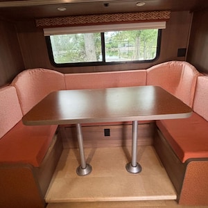 May include: Interior of a recreational vehicle with a booth-style seating area. The seating features coral-colored cushions and wooden frames. A rectangular table with a brown surface and silver legs is centered in the booth. A window with a view of trees is visible.