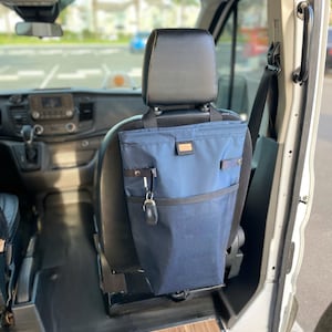 Puede incluir: Una bolsa de almacenamiento de tela azul cuelga de la parte posterior de un asiento de coche. La bolsa tiene un bolsillo con cremallera y un llavero unido a ella.