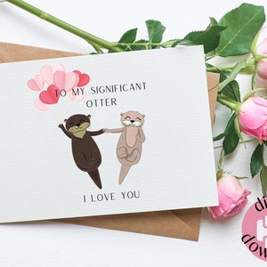 Könnte beinhalten: Eine Grußkarte mit dem Text "TO MY SIGNIFICANT OTTER I LOVE YOU" und Illustrationen von zwei Ottern, die sich an den Händen halten. Die Karte liegt auf einer weißen Oberfläche mit rosa Rosen und einem braunen Umschlag.