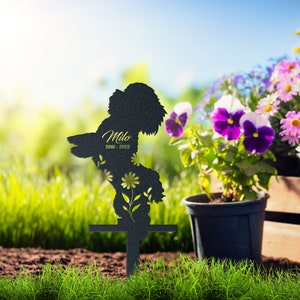 May include: A black metal garden stake in the shape of a dog, with the name "Milo" and dates "2010-2022" in a script font. The dog silhouette has floral cutouts. The stake is placed in a garden with green grass and colorful flowers.