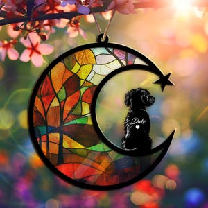 May include: A stained glass moon ornament with a silhouette of a dog inside. The moon has a colorful stained glass design with trees and a landscape. The dog is black and has the name "Duke" written on it.