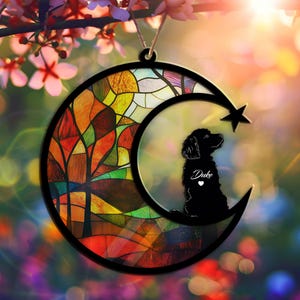 May include: A stained glass moon ornament with a silhouette of a dog and the name "Duke" inside. The moon has a colorful stained glass design with a forest scene.