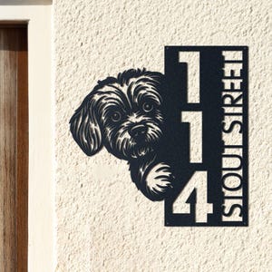 May include: Black metal address sign featuring a dog peeking out from behind the numbers "114". The word "STOUT STREET" is vertically displayed on the right side of the sign. The sign is mounted on a textured, off-white wall.