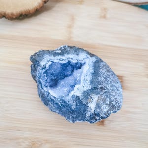 Peut inclure: Une géode bleue avec un centre rempli de cristaux, posée sur une surface en bois clair. L'extérieur de la géode est gris foncé avec des accents blancs, et les cristaux intérieurs sont d'une nuance de bleu plus claire.