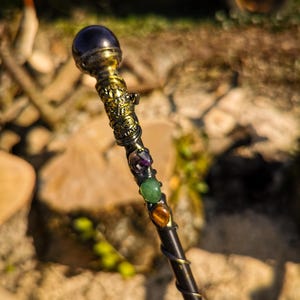 Peut inclure: Une baguette artisanale avec un manche en bois foncé et des détails en laiton ornés. La baguette présente une boule de cristal violet foncé au sommet, avec des pierres précieuses colorées et un motif en spirale. La baguette mesure environ 30 cm de long.