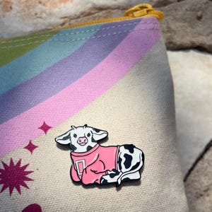May include: A white and black cow wearing a pink sweater with a white letter D on the front. The cow is sitting on a rainbow patterned fabric.