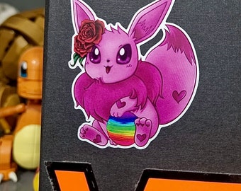 Op de afbeelding: Een sticker met een roze cartoonfiguur met grote ogen, een roos en een pluizige staart. Het figuur houdt een regenboogkleurige bal vast en heeft hartvormige details. De sticker bevindt zich op een zwart oppervlak.