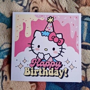 May include: A square birthday card with a cartoon cat wearing a party hat and bow. The card has a pink and white background with the words "Happy Birthday!" in a retro font. Decorative diamonds and plus signs surround the text.