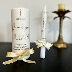 May include: Two white candles with gold lettering. The larger candle has the text "God Bless Penelope Liliani Baptised on 26th of June 2023 St Albert's Cathedral". The smaller candle has the text "Penelope Liliani Baptised 26/07/13".
