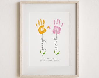 Puede incluir: Una impresión enmarcada con dos huellas de manos en amarillo y rosa, formando flores con los nombres "George" y "Sarah" escritos en los tallos. El texto "¡Manos arriba para el mejor papá del mundo!" está debajo de las huellas de manos.