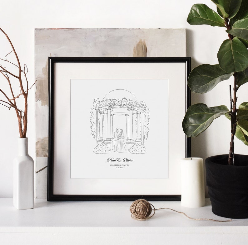 May include: Black framed line art print of a wedding scene. The print features a couple standing under an arbor, with the names "Paul & Olivia" and "Alberton Chapel" below. A white vase with branches, a candle, and a potted plant are also visible.