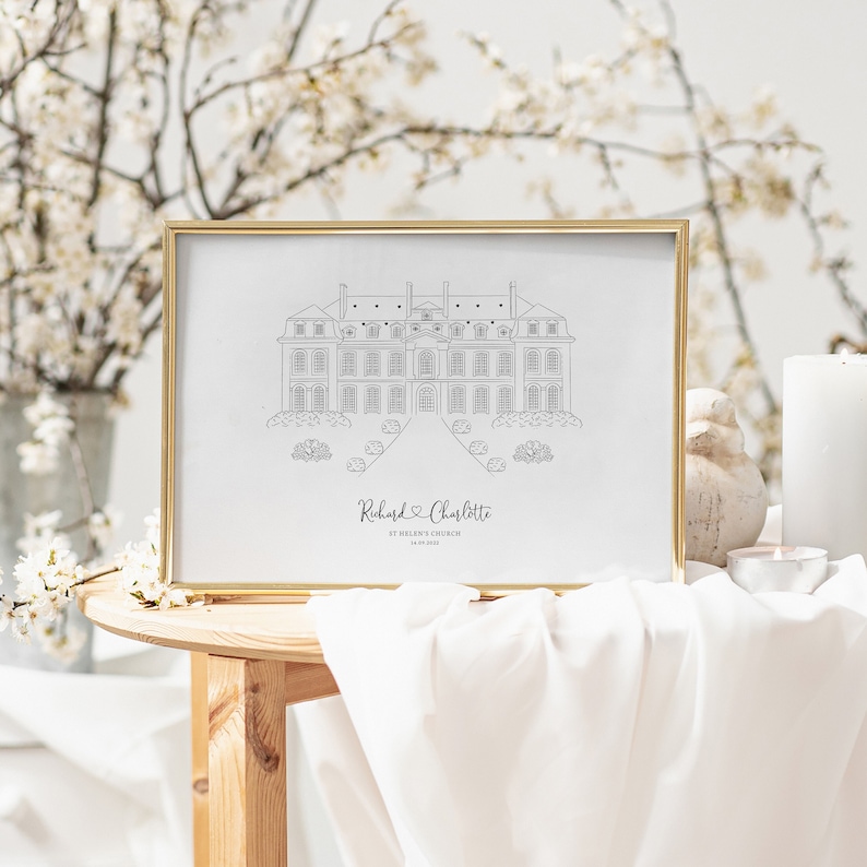 May include: A framed black and white line drawing of a stately building, likely a church or manor house, with the names "Richard & Charlotte" and a date below. The gold frame sits on a wooden table with white fabric.