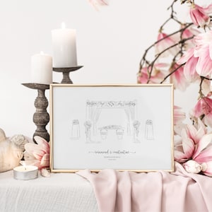 May include: A framed line drawing of a wedding ceremony scene, featuring a floral arch and table, with the names "Rosamond & Constantine" and the date below. The frame is gold, and the image is set against a backdrop of candles and pink flowers.