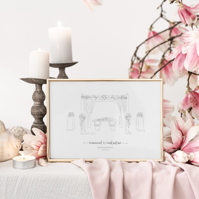 May include: A framed line drawing of a wedding ceremony scene, featuring a floral arch and table, with the names "Rosamond & Constantine" and the date below. The frame is gold, and the image is set against a backdrop of candles and pink flowers.