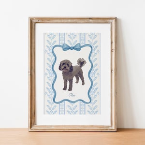 May include: A framed print featuring a watercolor illustration of a dog named Thea. The dog is depicted in a standing pose within a blue frame with a bow at the top. The background has a blue and white floral pattern.