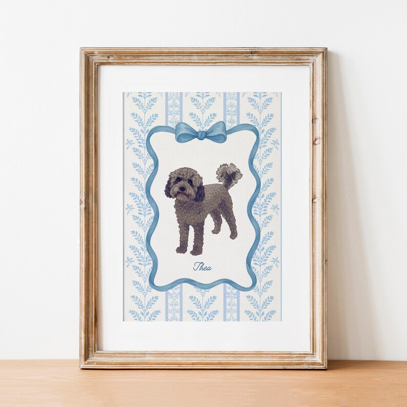 May include: A framed print featuring a watercolor illustration of a dog named Thea. The dog is depicted in a standing pose within a blue frame with a bow at the top. The background has a blue and white floral pattern.