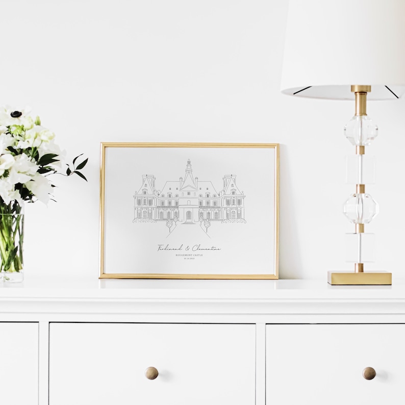 May include: A framed print featuring a line drawing of a castle, displayed on a white dresser. The artwork is in a gold frame and includes the text "Ferdinand & Clementine" and "Rosemont Castle". A white lamp and a vase of white flowers are also visible.