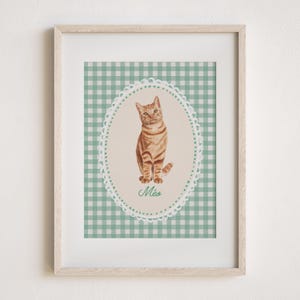 May include: Framed print featuring a watercolor illustration of a tabby cat. The cat is inside an oval frame with a scalloped edge, set against a green and white gingham background. The word "M&eacute;a" is written in script below the cat.
