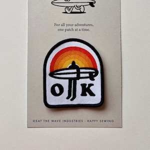 May include: A white iron-on patch with a black and white illustration of a surfer riding a wave with a sunset behind them. The text "OK" is written in black below the surfer. The text "EAT THE WAVE INDUSTRIES - HAPPY SEWING" is printed below the patch.