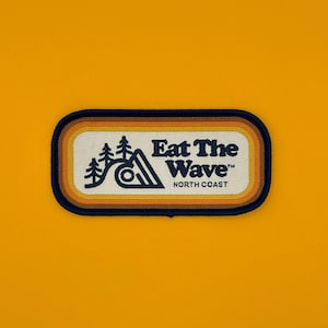 Op de afbeelding: Een geborduurde patch in geel, oranje en bruin met de tekst "Eat The Wave" in donkerblauwe letters. De tekst "North Coast" staat ook in donkerblauwe letters onder de hoofdtekst. De patch heeft een gestileerde afbeelding van een golf met bomen op de achtergrond.