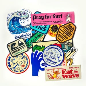 Op de afbeelding: Een verzameling kleurrijke stickers met surfthema's. Ontwerpen omvatten een cartoon golf, een biddende hand en tekst die luidt "Pray for Surf" en "Eat the Wave".