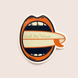 Könnte beinhalten: Orangefarbener und weißer Aufkleber mit einem Mund mit Zähnen, der in ein Surfbrett beißt, mit dem Text "Eat the Wave".