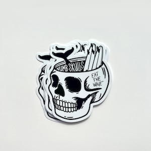 Può includere: Adesivo in bianco e nero di un teschio con una coda di balena e tavole da surf all'interno. Il testo "Numb Skulls" e "Eat the Wave" sono sull'adesivo.