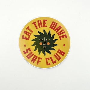 May include: A circular yellow sticker featuring red text and a black sun design. The text says "EAT THE WAVE . SURF CLUB." The sun has closed eyes, rosy cheeks, and a small mouth. The sticker has a white border.