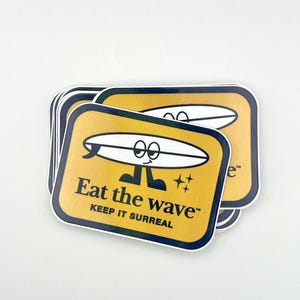 May include: A stack of square stickers with a yellow background and a white surfboard graphic with a face. The text "Eat the wave" and "Keep it surreal" are printed in black. The stickers have a navy blue border.