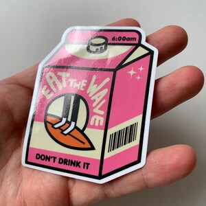 May include: Pink milk carton sticker featuring the text 'Eat the Wave' and 'Don't Drink It'.  The design includes a cartoon surfer and a barcode. The time '6:00am' is printed on the top of the carton. The sticker has a glossy finish.