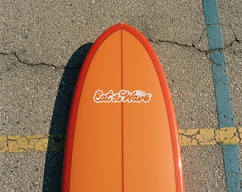 Könnte beinhalten: Ein orangefarbenes Surfbrett mit rotem Rand und dem Text "Eat the Wave" in weißer, stilisierter Schrift. Das Surfbrett liegt auf einer strukturierten grauen Oberfläche mit gelben und blauen Linien.