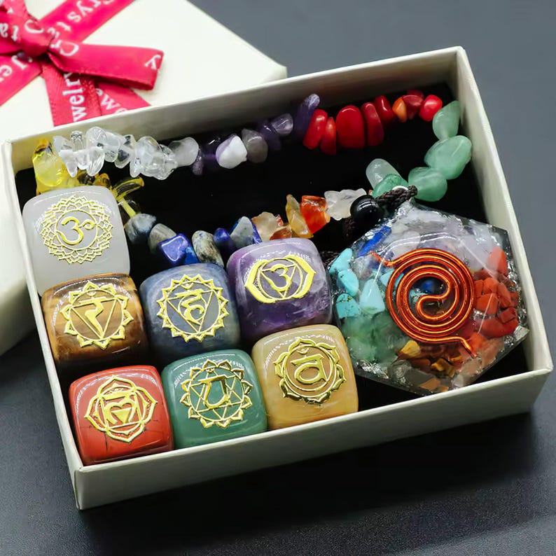 K&ouml;nnte beinhalten: Ein Set aus sieben Chakra-Steinen in einer wei&szlig;en Box. Jeder Stein hat eine andere Farbe und ein goldenes Symbol. Die Steine sind in einem Kreis angeordnet, mit einem gr&ouml;&szlig;eren Stein in der Mitte. Die Steine sind von kleineren Steinen in verschiedenen Farben umgeben.