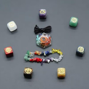 K&ouml;nnte beinhalten: Ein Set aus sieben Chakra-Steinen, jeder mit einer anderen Farbe und einem anderen Symbol, angeordnet in einem Kreis um einen Chakra-Anh&auml;nger. Die Steine sind klein und quadratisch, und der Anh&auml;nger hat eine sechseckige Form mit einem Spiralmuster. In der Mitte des Kreises befindet sich auch eine Schnur mit bunten Perlen.