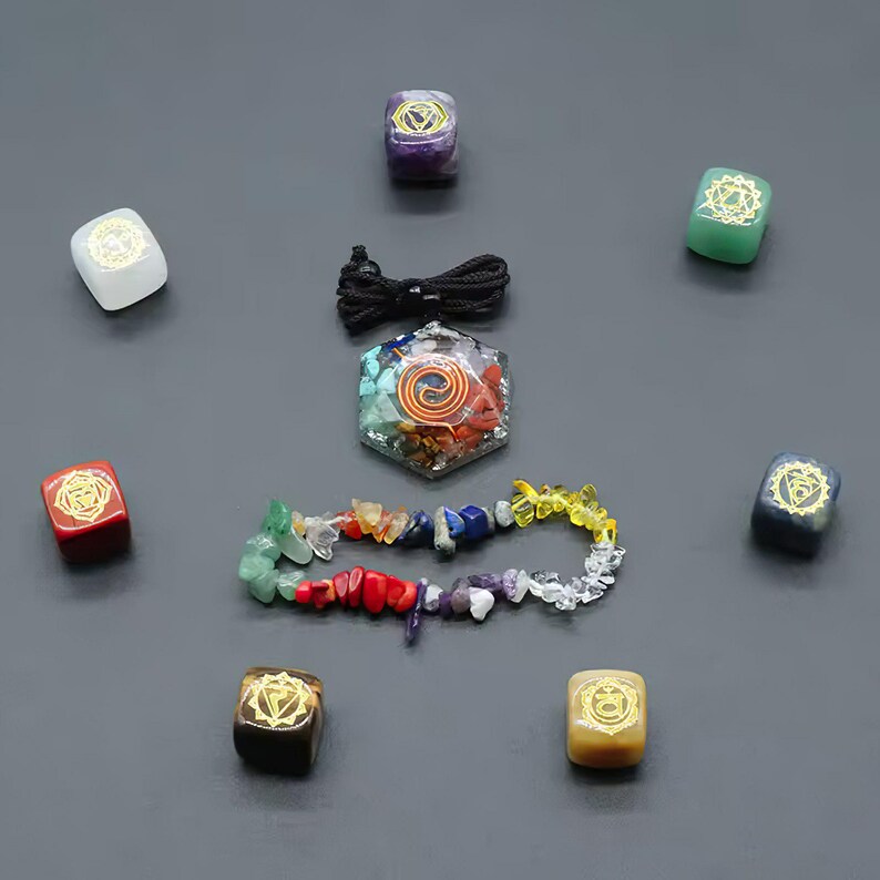 K&ouml;nnte beinhalten: Ein Set aus sieben Chakra-Steinen, jeder mit einer anderen Farbe und einem anderen Symbol, angeordnet in einem Kreis um einen Chakra-Anh&auml;nger. Die Steine sind klein und quadratisch, und der Anh&auml;nger hat eine sechseckige Form mit einem Spiralmuster. In der Mitte des Kreises befindet sich auch eine Schnur mit bunten Perlen.