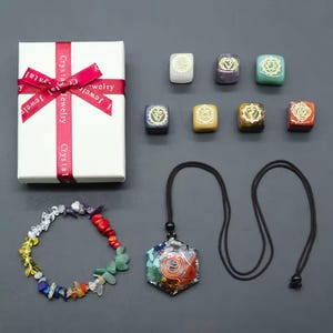 K&ouml;nnte beinhalten: Sieben Chakra-Steine in kleinen Schachteln, ein Armband mit bunten Steinen und eine Halskette mit einem sieben Chakra-Stein-Anh&auml;nger. Die Steine sind in den Farben der Chakren: Rot, Orange, Gelb, Gr&uuml;n, Blau, Indigo und Violett. Die Halskette hat eine schwarze Schnur und einen sechseckigen Anh&auml;nger mit einem Spiralmuster.