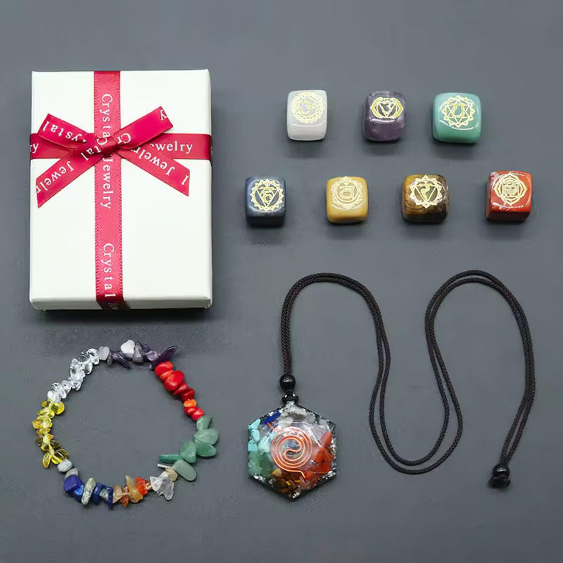 K&ouml;nnte beinhalten: Sieben Chakra-Steine in kleinen Schachteln, ein Armband mit bunten Steinen und eine Halskette mit einem sieben Chakra-Stein-Anh&auml;nger. Die Steine sind in den Farben der Chakren: Rot, Orange, Gelb, Gr&uuml;n, Blau, Indigo und Violett. Die Halskette hat eine schwarze Schnur und einen sechseckigen Anh&auml;nger mit einem Spiralmuster.