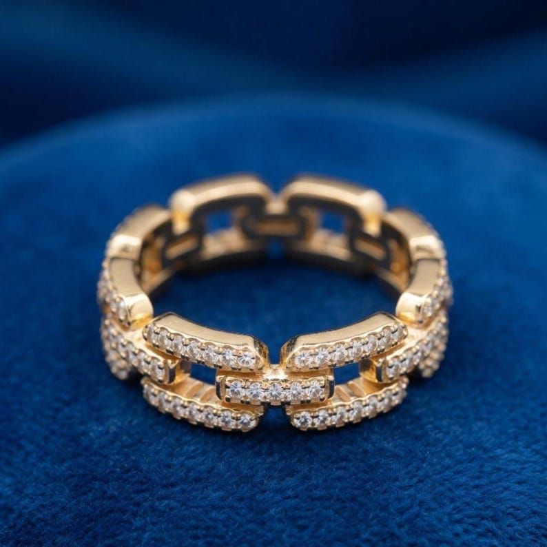 May include: A gold ring featuring a chain link design, adorned with small, clear gemstones. The ring is set against a dark blue backdrop. The ring's design is intricate and eye-catching.