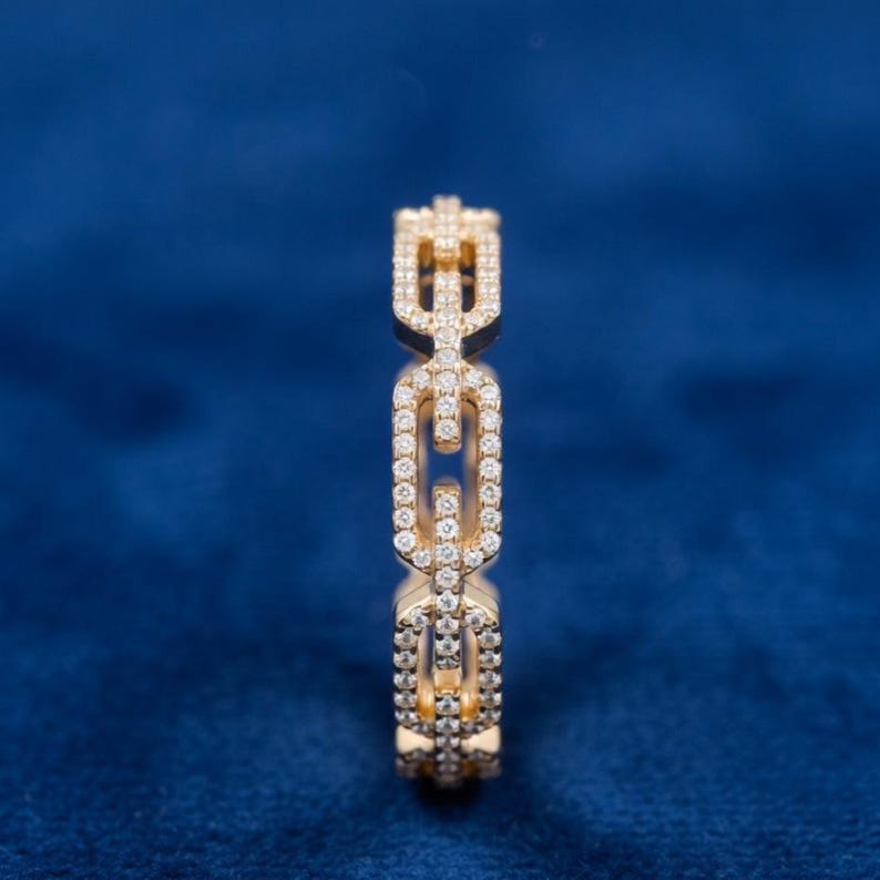 May include: A gold ring featuring a chain link design, studded with small, clear gemstones. The ring's links are detailed and the gemstones sparkle. The background is a deep blue.