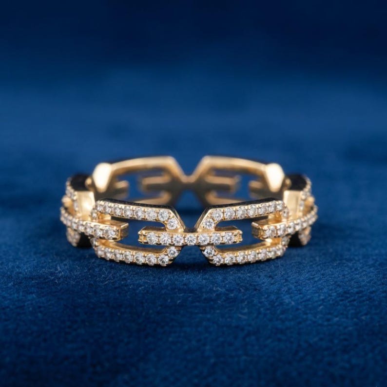 May include: A gold ring featuring a chain link design, adorned with small diamonds. The ring is set against a dark blue backdrop, accentuating its intricate details and luxurious appearance. The ring's design is modern and elegant, suitable for various occasions.